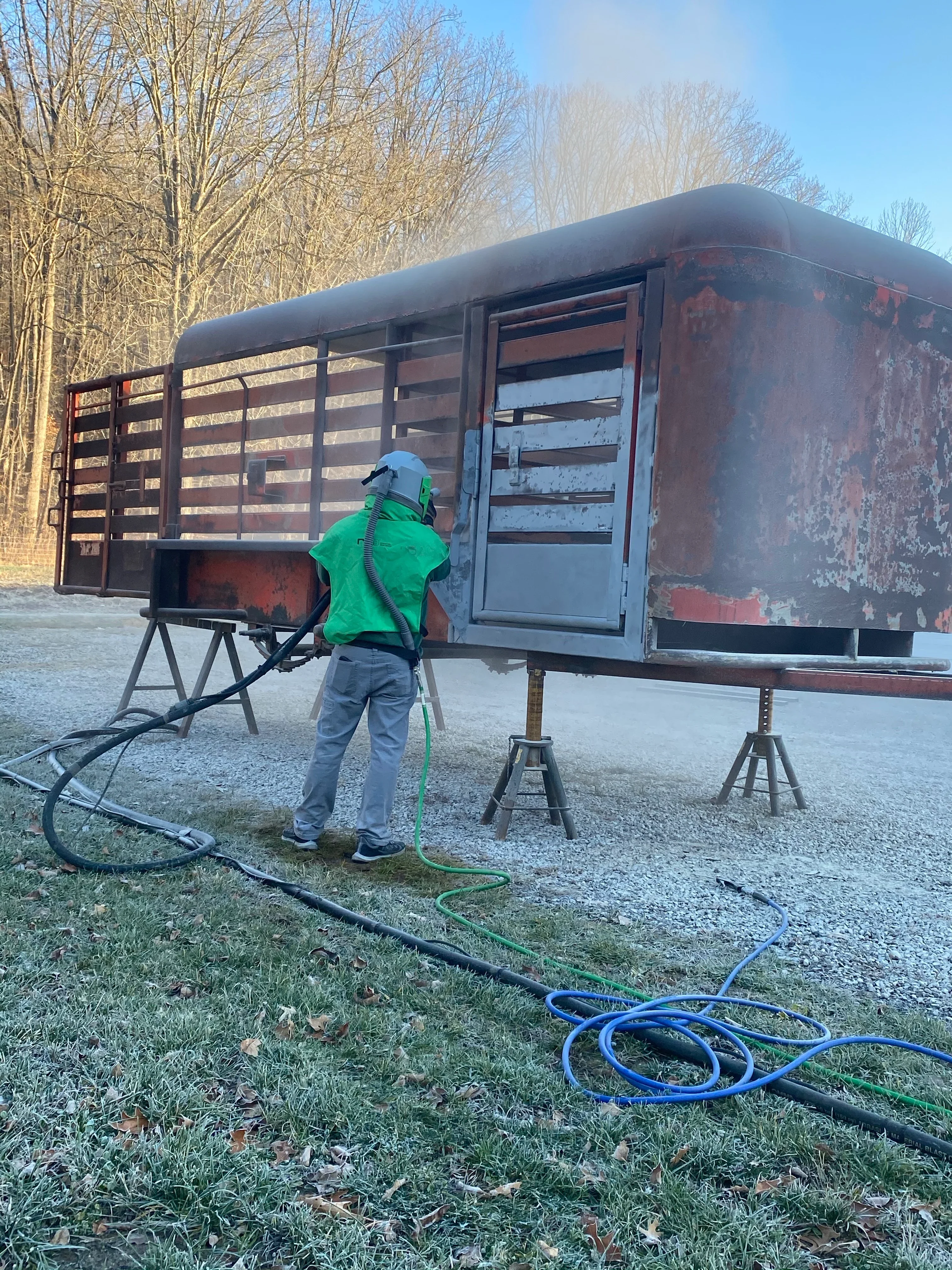 Worker sandblasting rusted steel horse trailer with pressure equipment and hoses